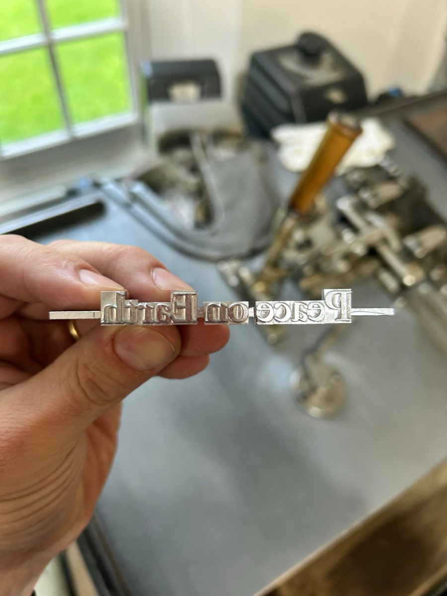 A freshly cast metal type slug reading Peace on Earth, held above the caster